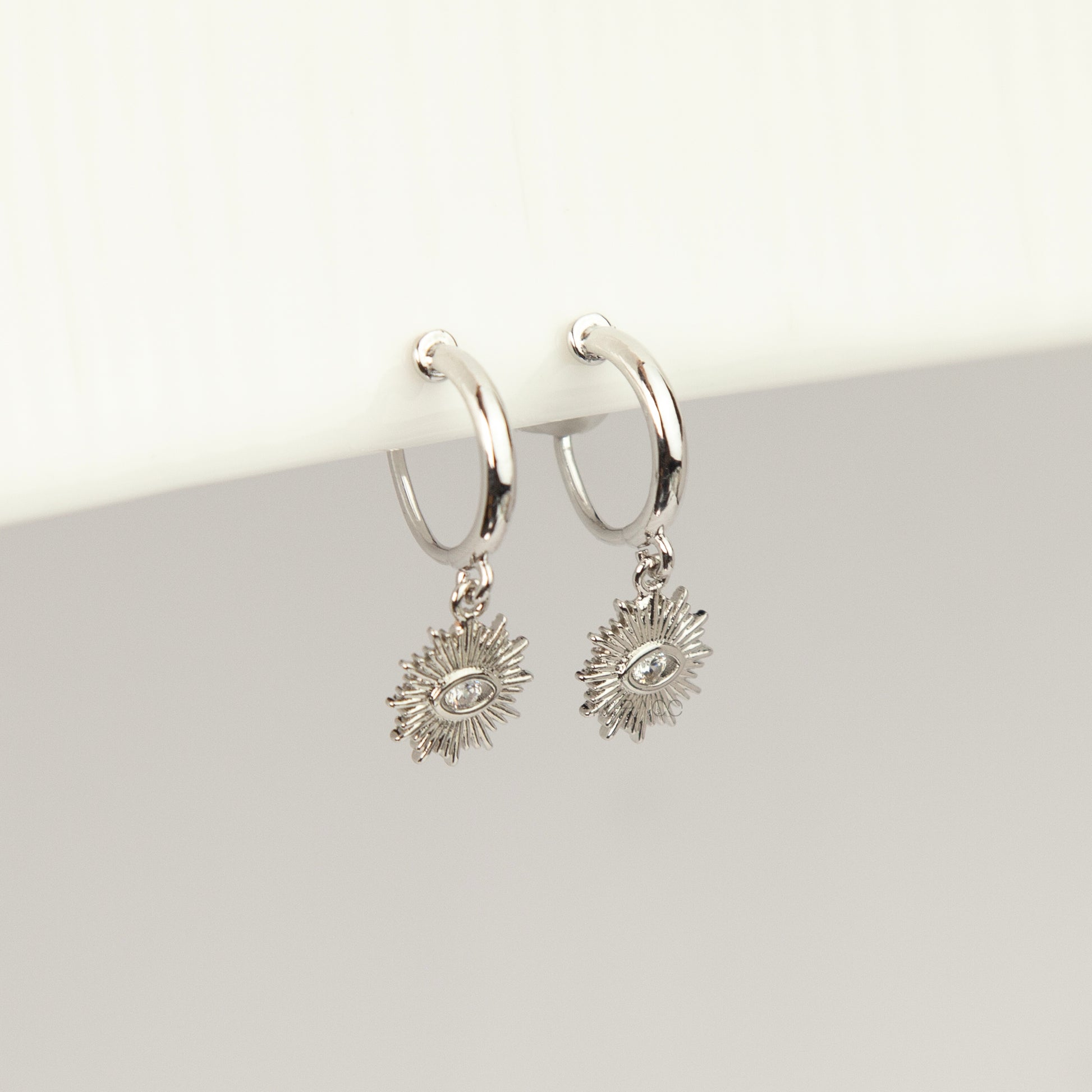 Silver coil-back clip-on hoop earrings featuring a dangling sunburst charm with a central oval crystal evil eye motif 
