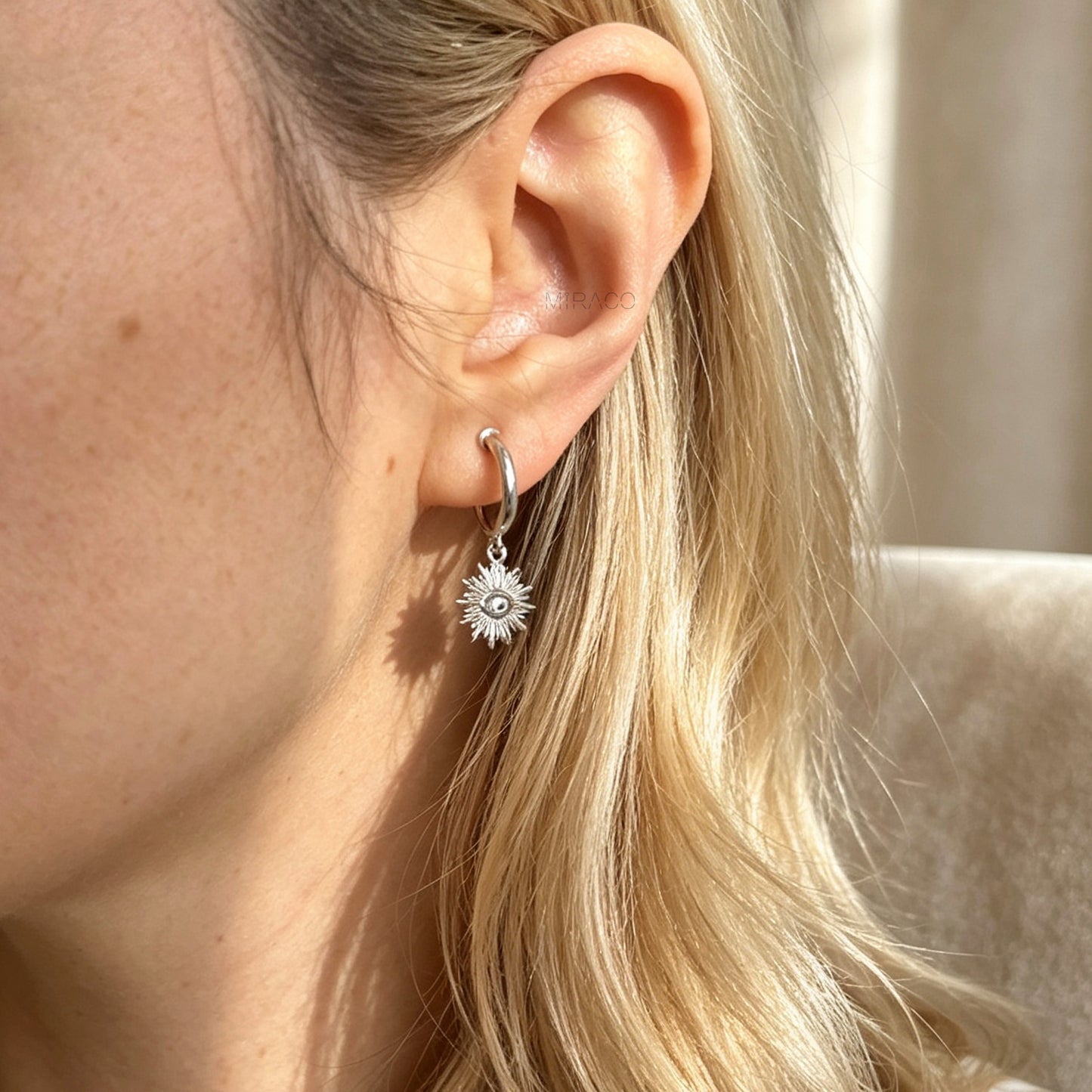 Silver coil-back clip-on hoop earrings featuring a dangling sunburst charm with a central oval crystal evil eye motif worn on a model.