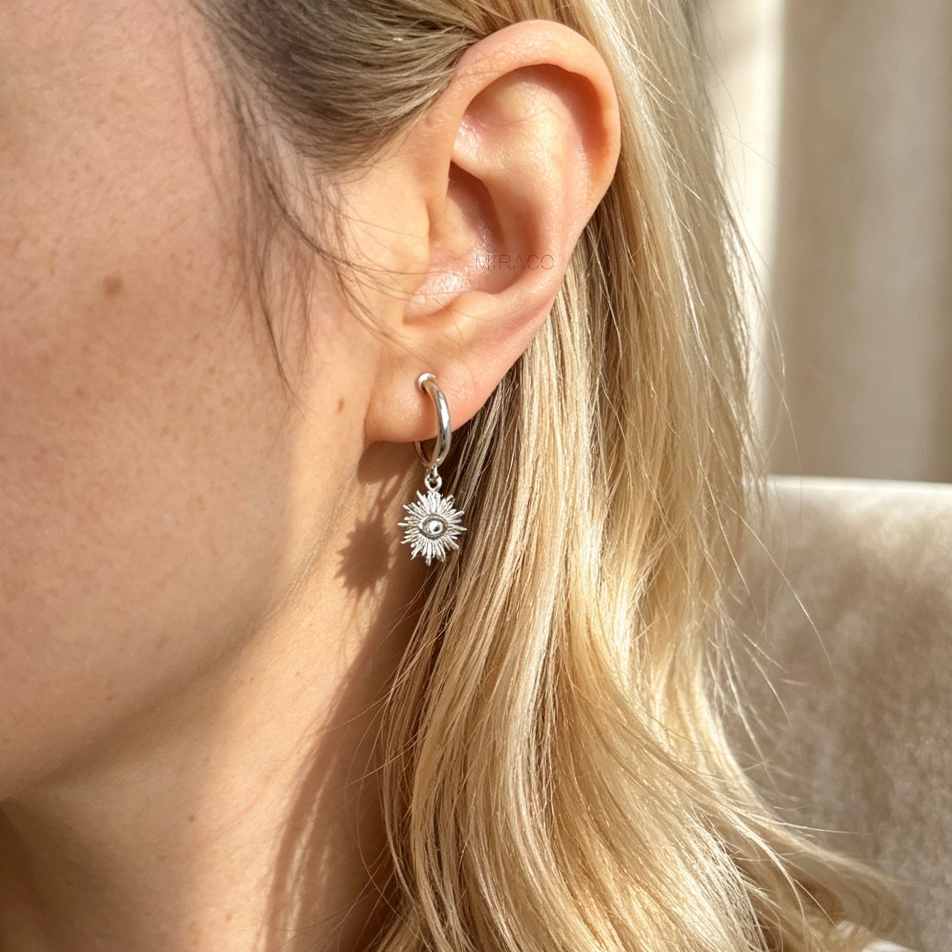 Silver coil-back clip-on hoop earrings featuring a dangling sunburst charm with a central oval crystal evil eye motif worn on a model.