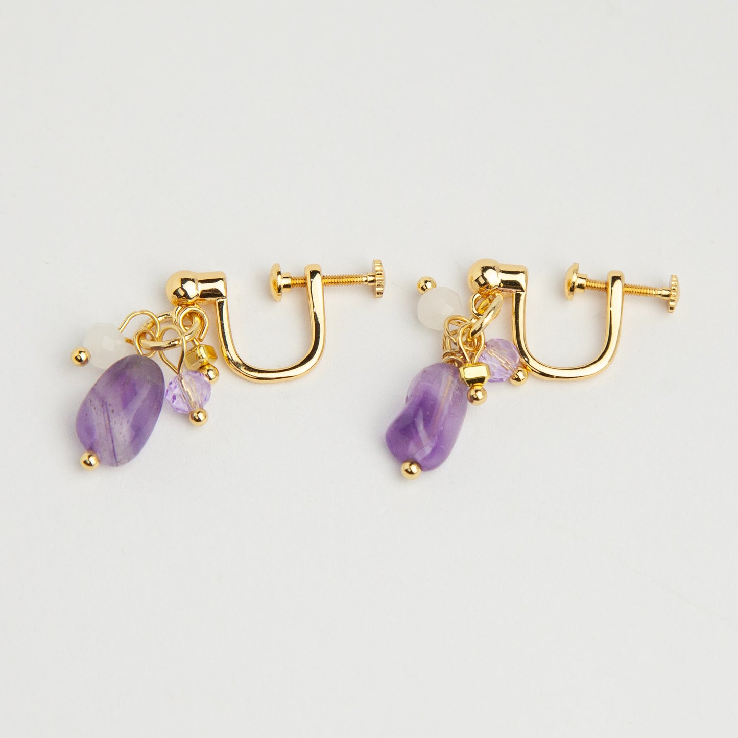 Gold screw-back clip-on hoop earrings with a raw purple amethyst stone, faceted lilac crystal charm, and white bead displayed on a light background.