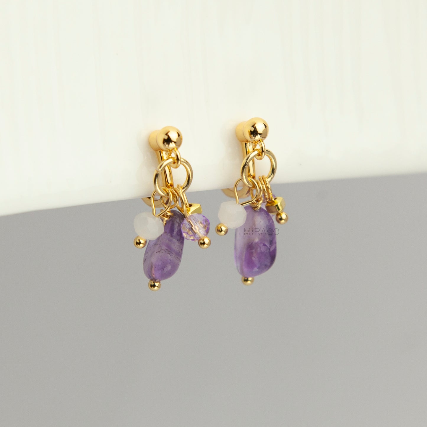 Gold screw-back clip-on hoop earrings with a raw purple amethyst stone, faceted lilac crystal charm, and white bead displayed on a light background.