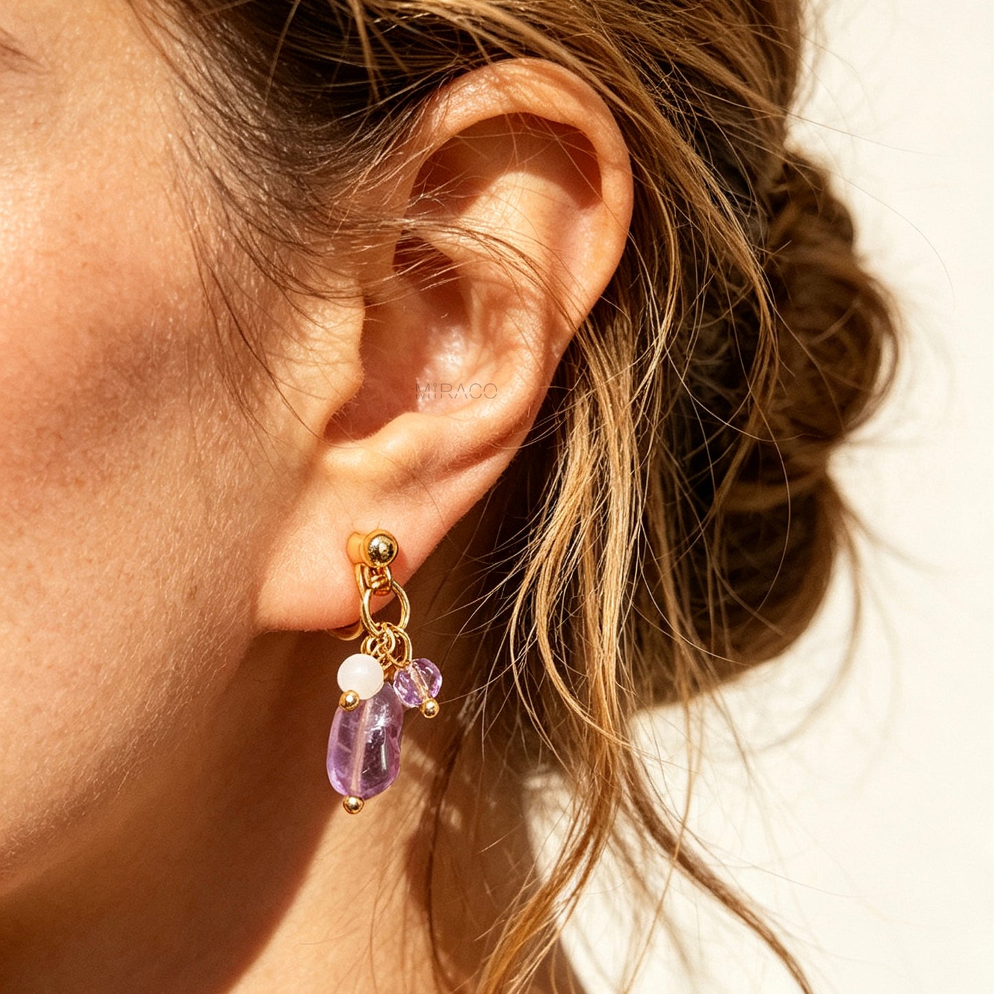 Gold screw-back clip-on hoop earrings with a raw purple amethyst stone, faceted lilac crystal charm, and white bead displayed on a light background.