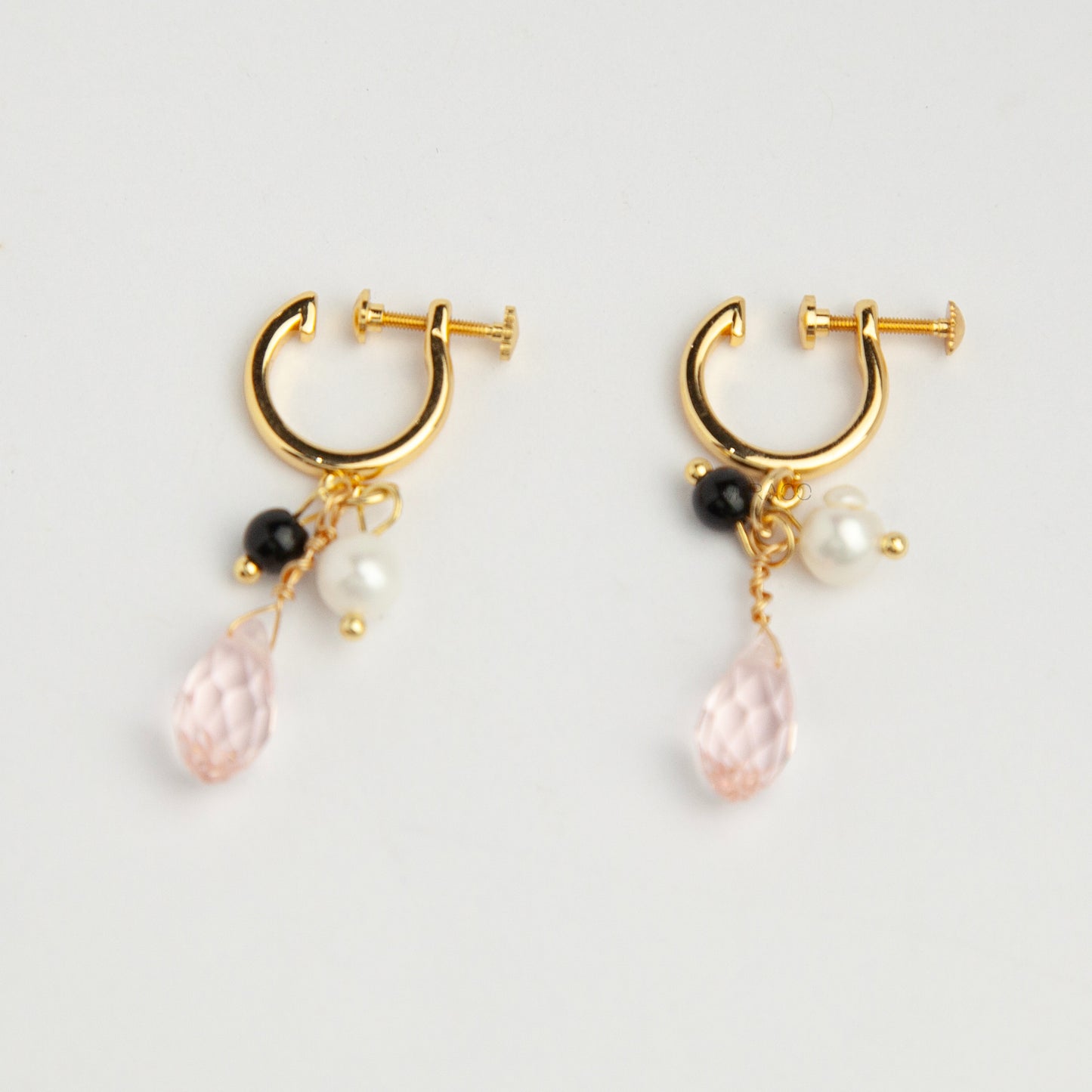 Gold screw-back clip-on hoop earrings featuring a faceted pink teardrop crystal charm, small white pearl, and black bead