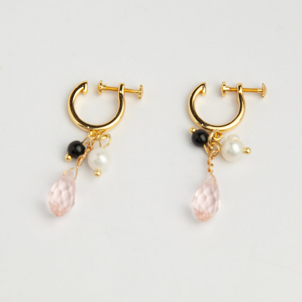 Gold screw-back clip-on hoop earrings featuring a faceted pink teardrop crystal charm, small white pearl, and black bead