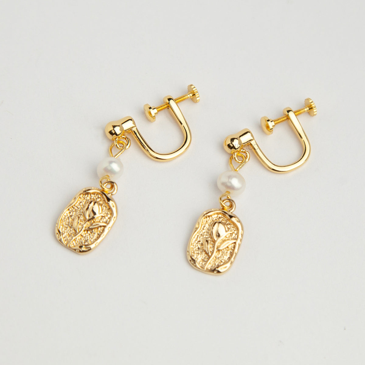Gold screw-back clip-on drop earrings featuring a lustrous white pearl and a textured oval medallion with an embossed floral design