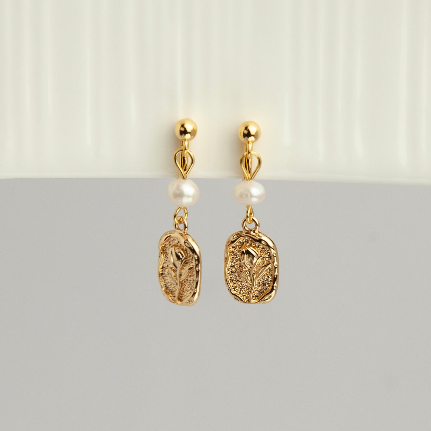 Gold screw-back clip-on drop earrings featuring a lustrous white pearl and a textured oval medallion with an embossed floral design