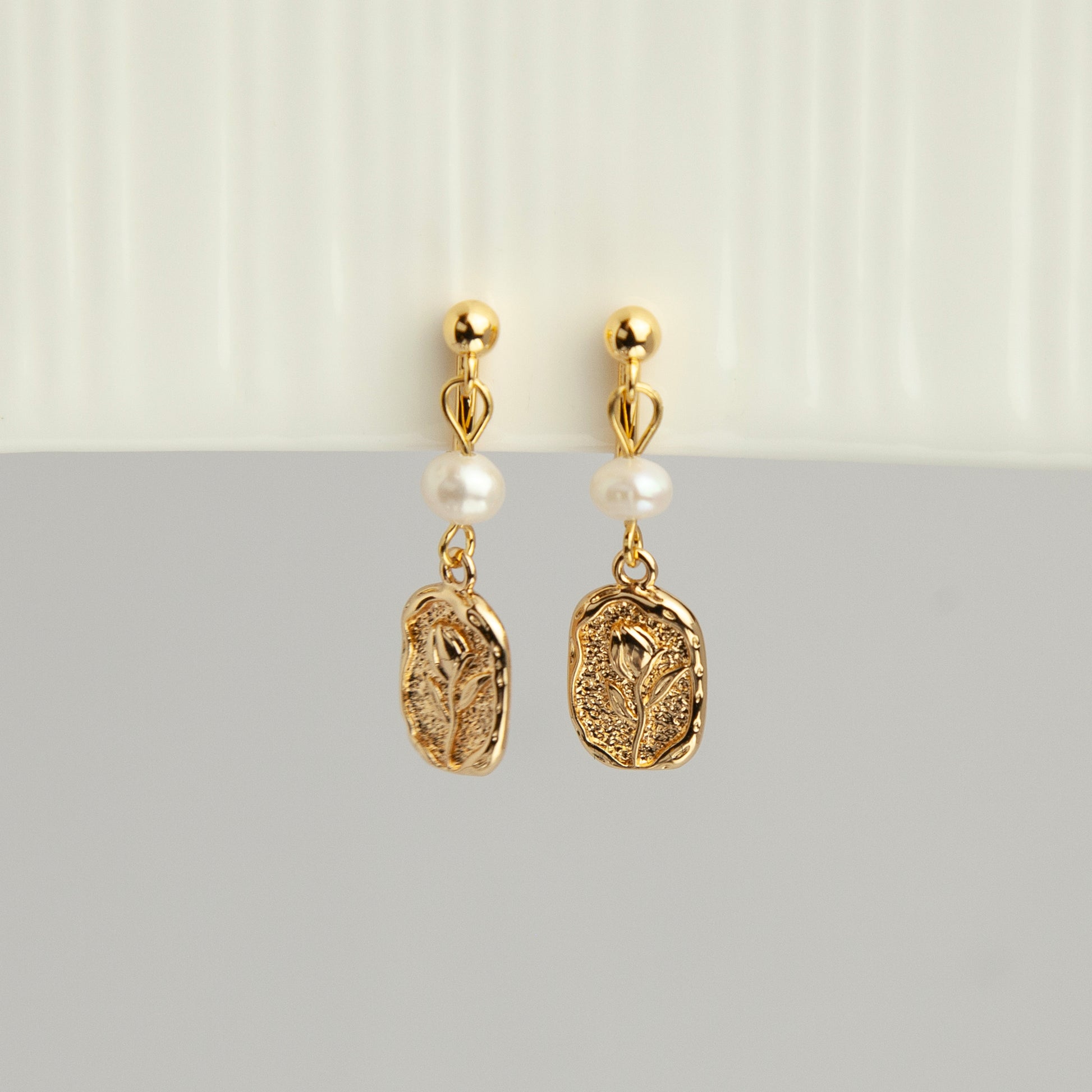Gold screw-back clip-on drop earrings featuring a lustrous white pearl and a textured oval medallion with an embossed floral design