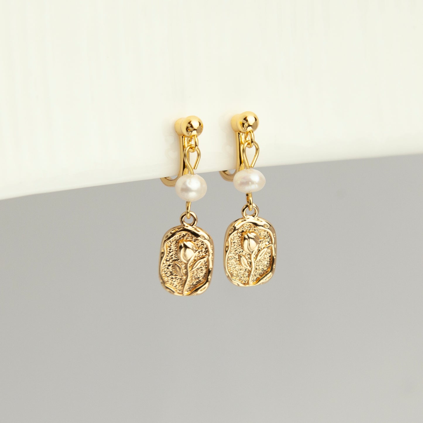 Gold screw-back clip-on drop earrings featuring a lustrous white pearl and a textured oval medallion with an embossed floral design