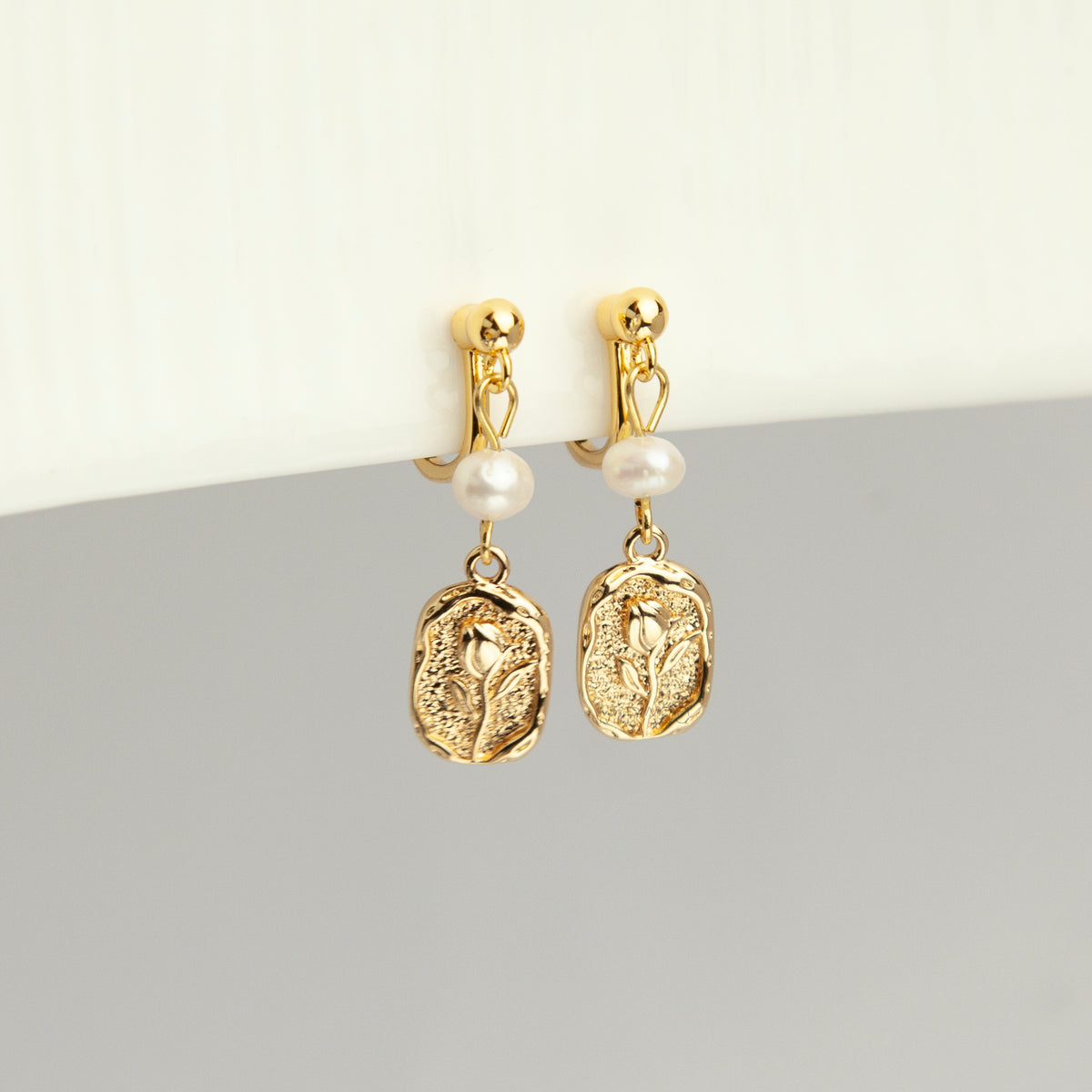Gold screw-back clip-on drop earrings featuring a lustrous white pearl and a textured oval medallion with an embossed floral design