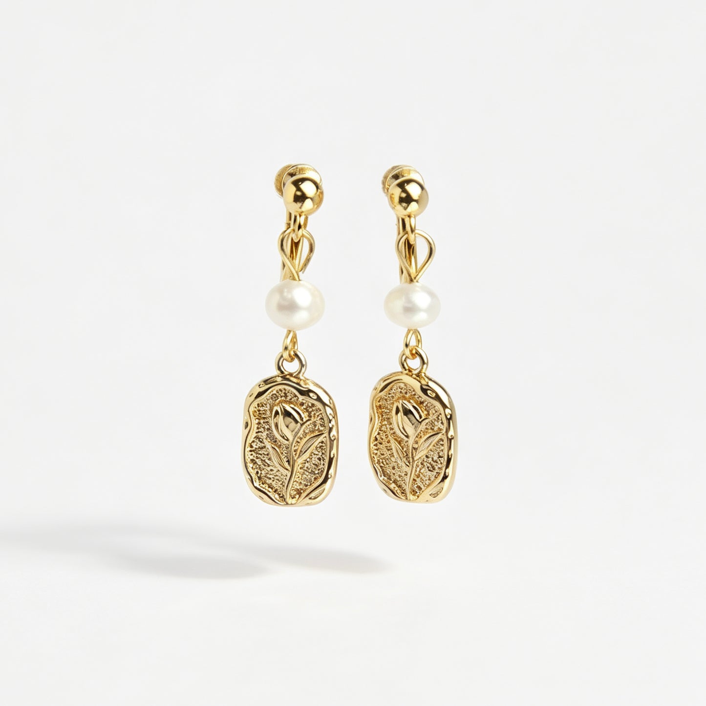 Gold screw-back clip-on drop earrings featuring a lustrous white pearl and a textured oval medallion with an embossed floral design