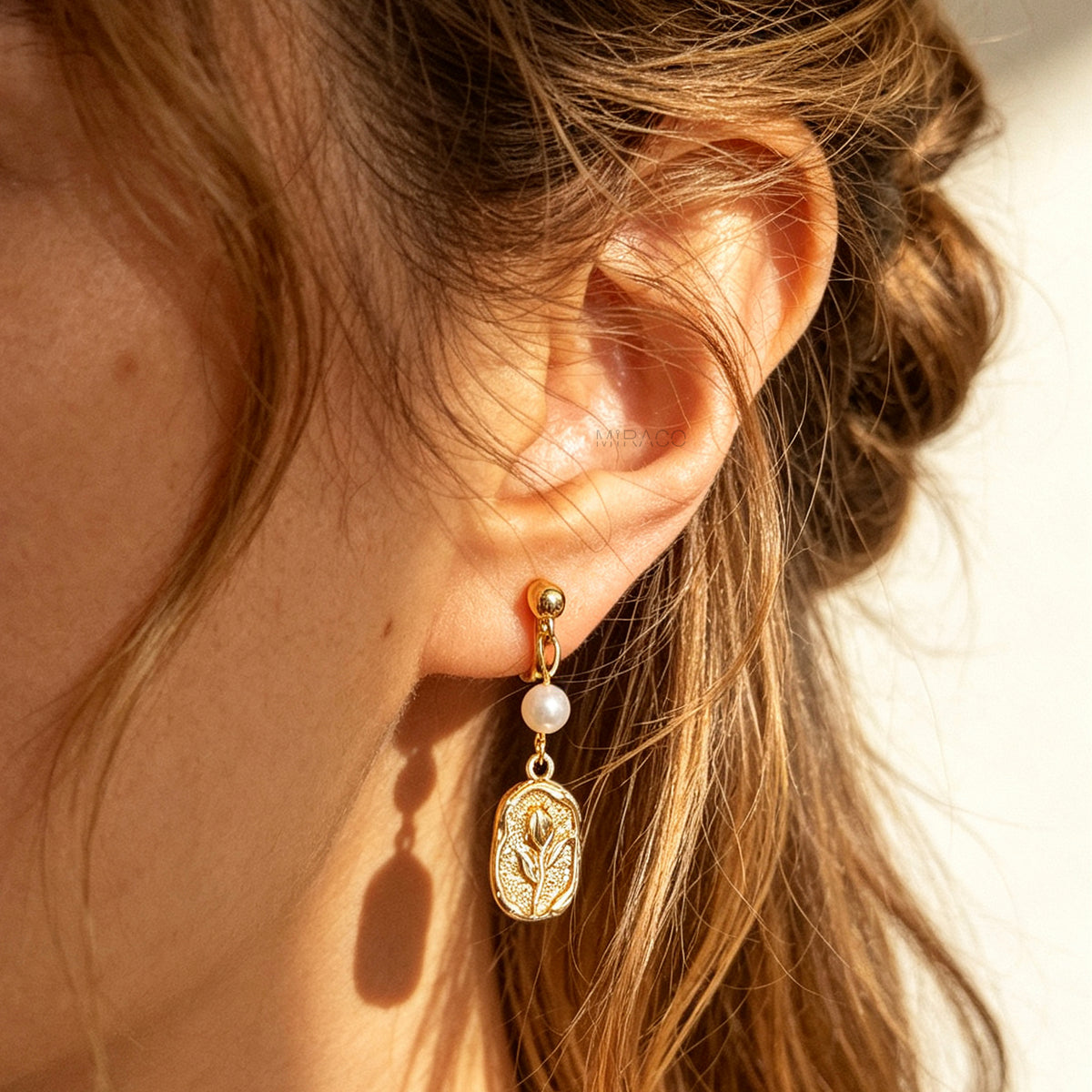 Gold screw-back clip-on drop earrings featuring a lustrous white pearl and a textured oval medallion with an embossed floral design