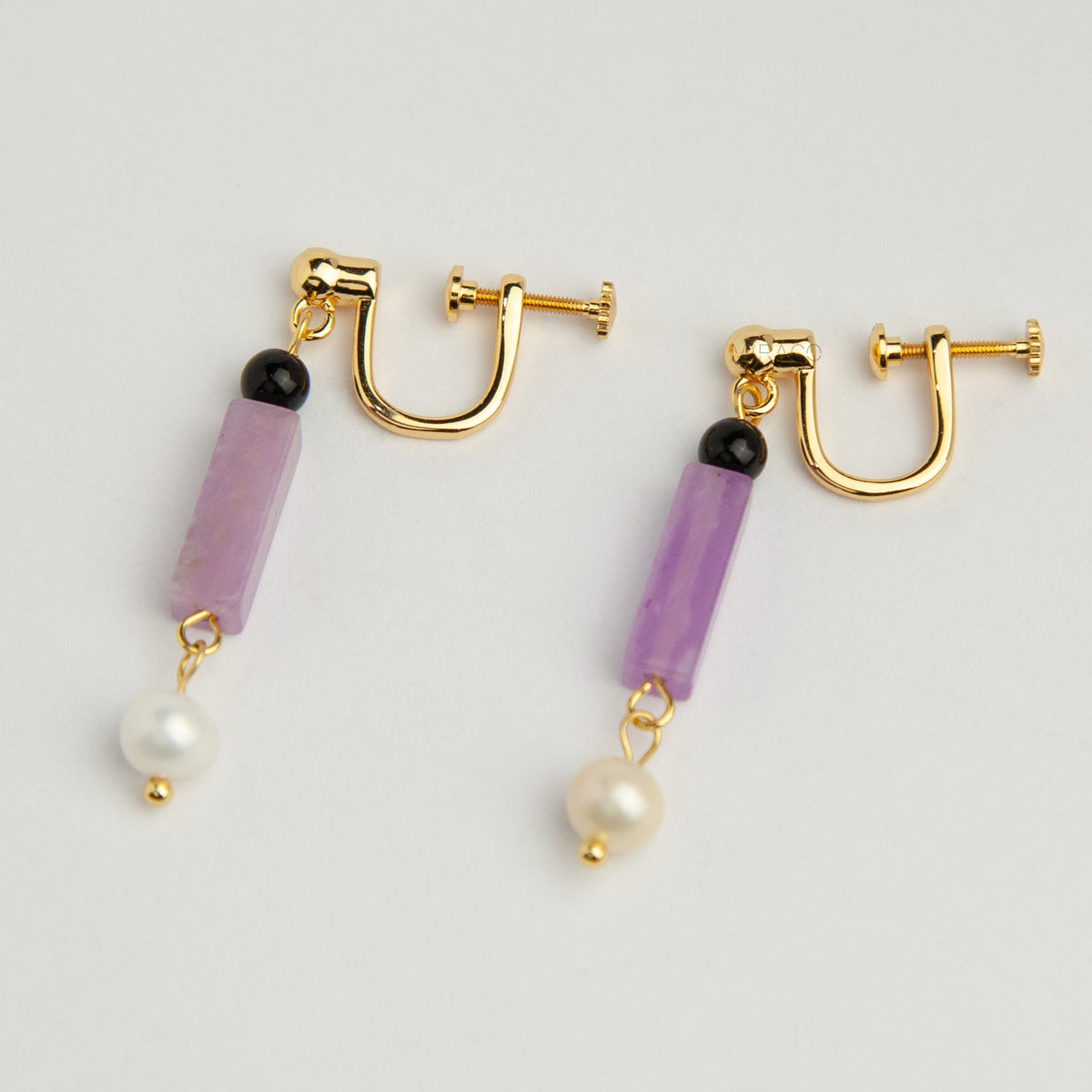 Gold screw-back clip-on drop earrings featuring a black onyx bead, rectangular purple jade stone, and dangling white freshwater pearl.