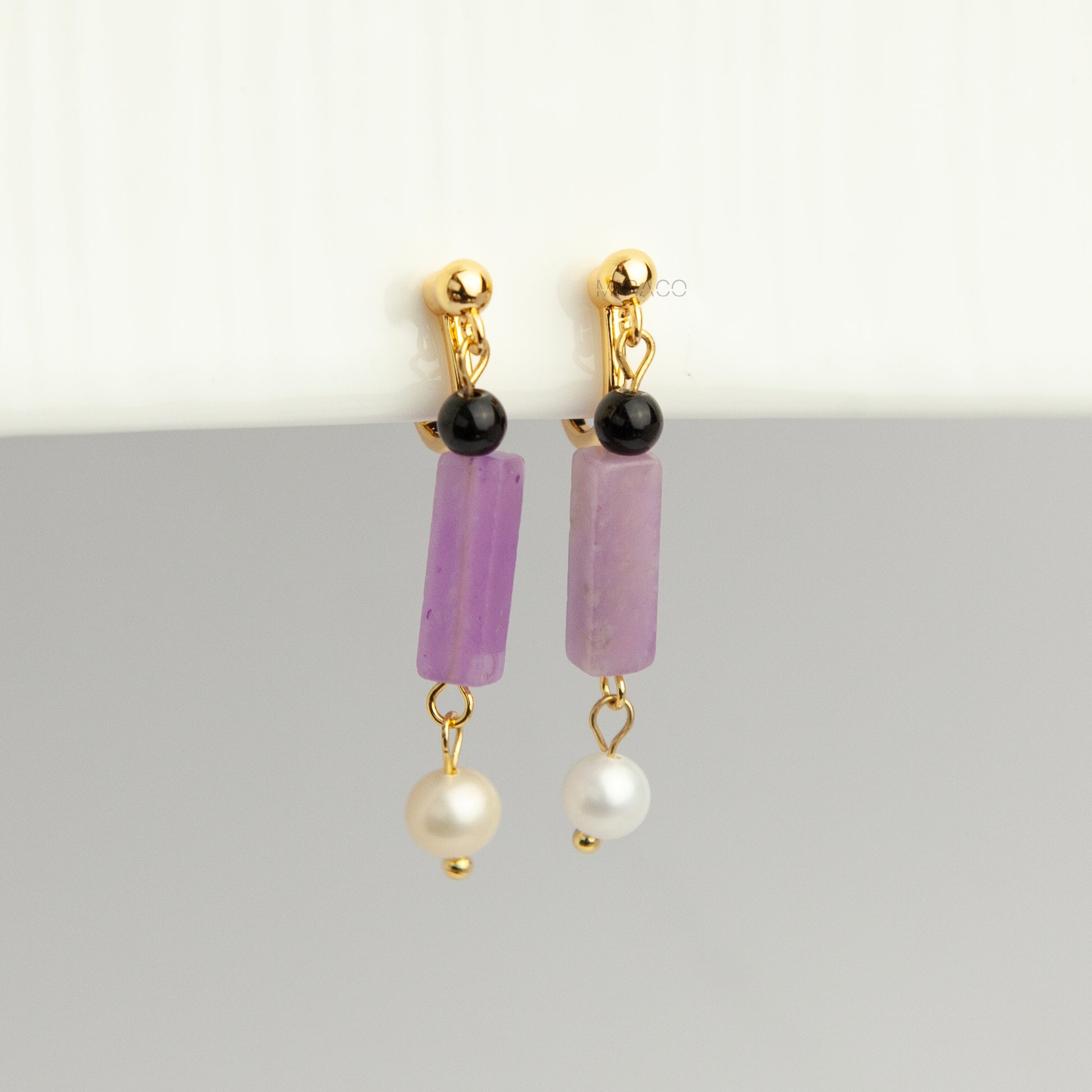 Gold screw-back clip-on drop earrings featuring a black onyx bead, rectangular purple jade stone, and dangling white freshwater pearl.