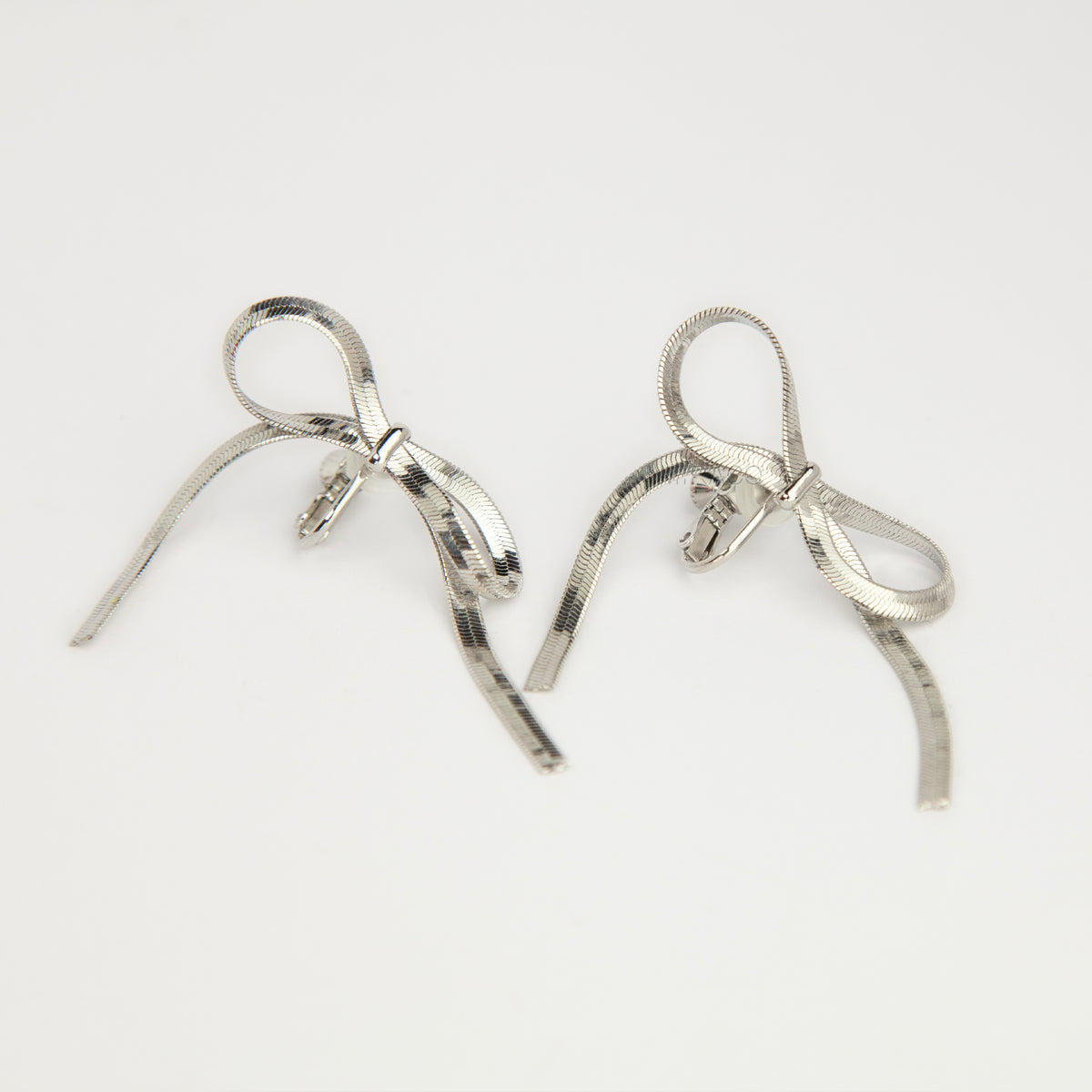 A pair of silver-toned dangling earrings shaped like ribbon bows, photographed on a plain white background.