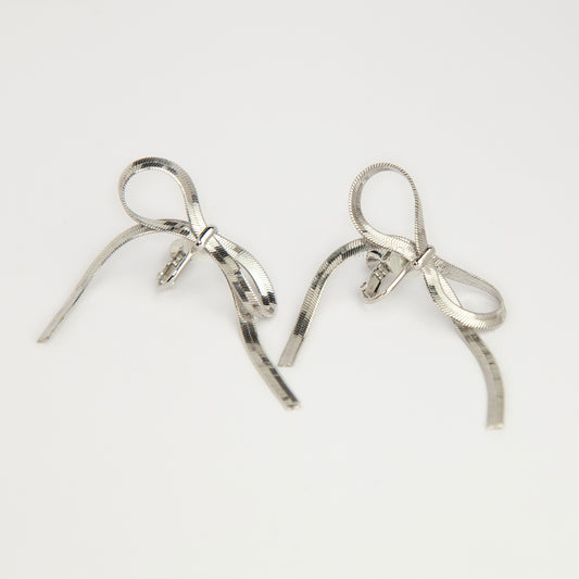 A pair of silver-toned dangling earrings shaped like ribbon bows, photographed on a plain white background.