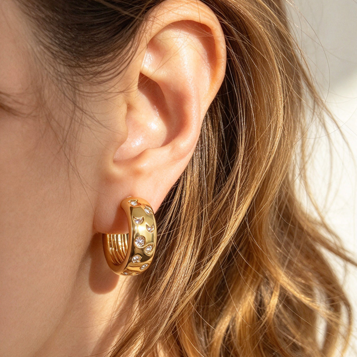 Woman wearing gold celestial star and moon cubic zirconia chunky hoop clip-on earrings with invisible clear resin backs for non-pierced ears.