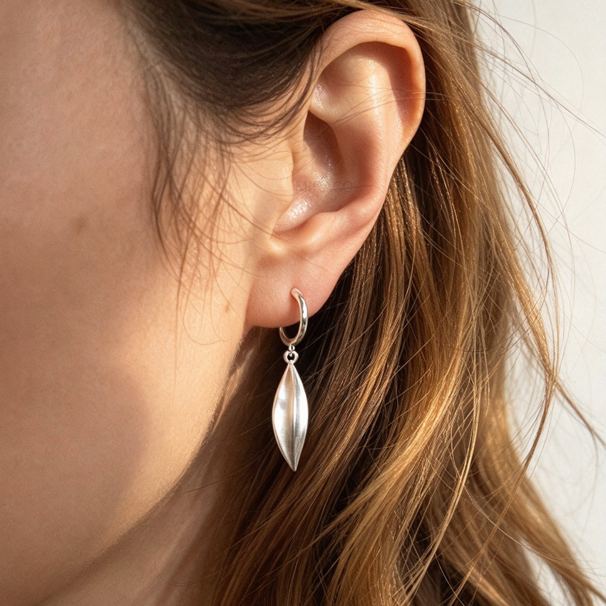 Woman wearing silver clip-on hoop earrings with brushed leaf drop pendants featuring a spring coil back for non-pierced ears.