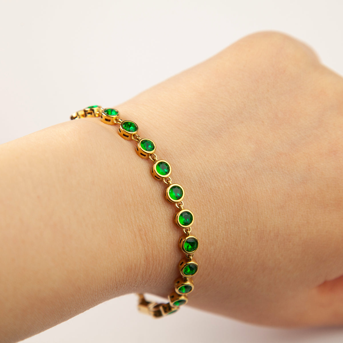 Woman wearing an 18K gold plated bezel-set emerald green cubic zirconia tennis bracelet on wrist, layered alongside a clear CZ stone version in waterproof stainless steel.