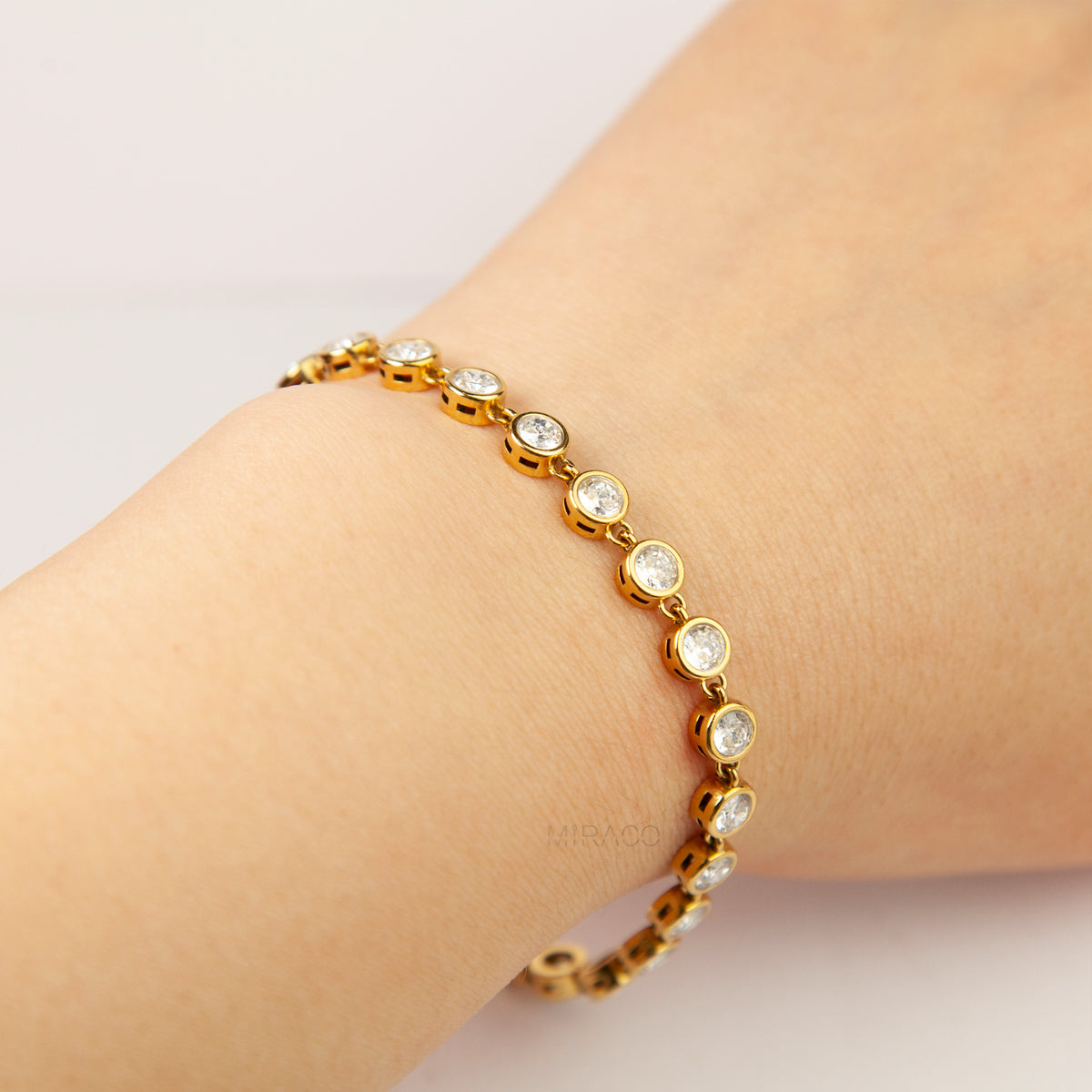 A delicate gold bracelet with round clear gemstones, shown worn on a person's wrist against a soft light background.