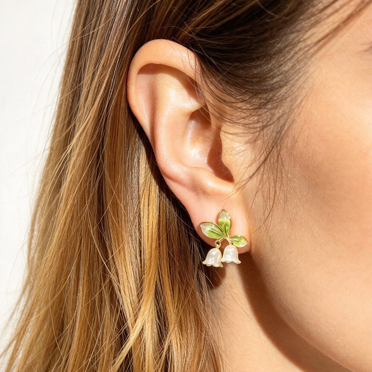 Woman wearing Lily of the Valley floral coil back clip-on earrings with green enamel leaves and pearlescent white flower drops in 18K gold plating on ear.