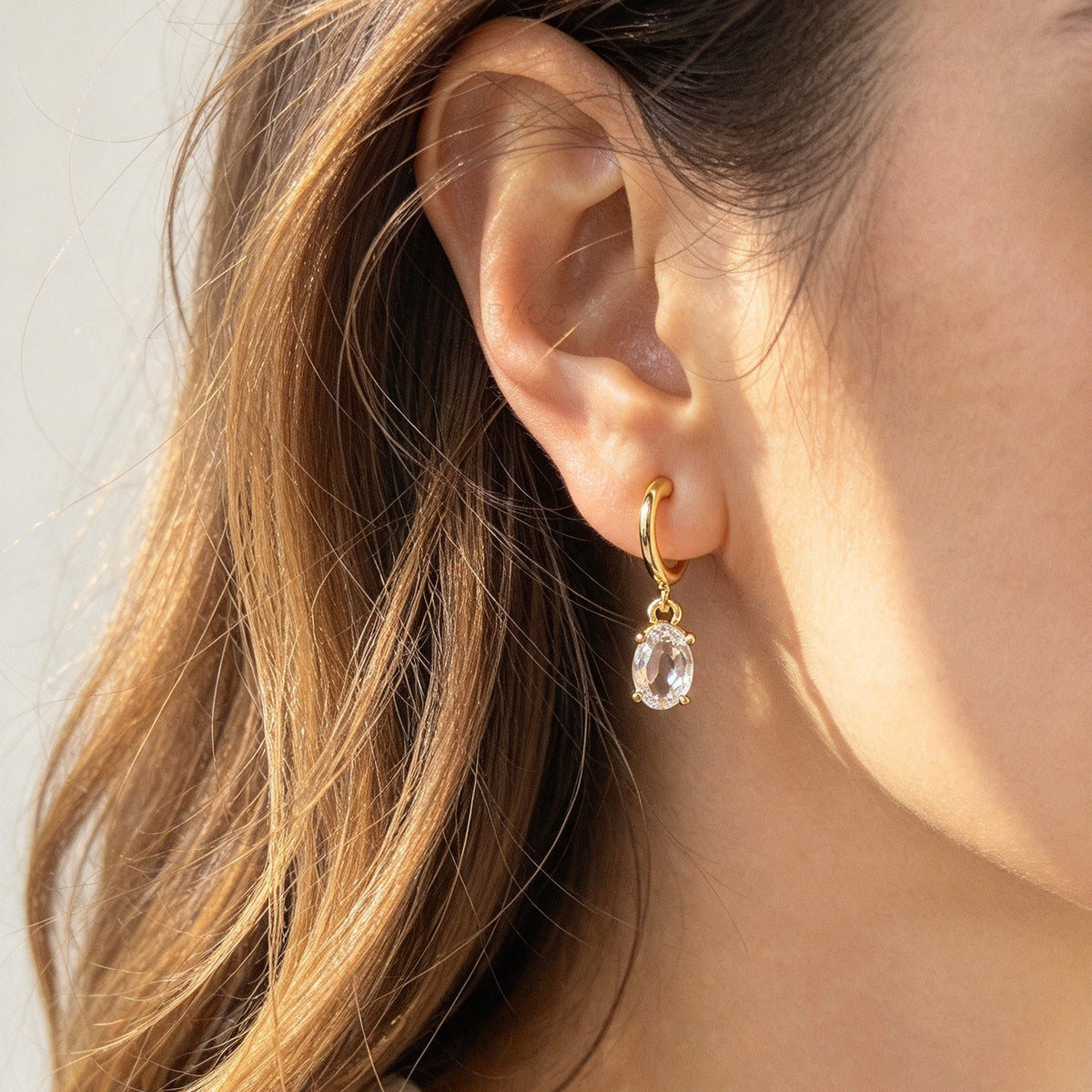Woman wearing 18K gold plated oval solitaire crystal huggie drop clip-on earrings with coil back non-pierced closure on ear.